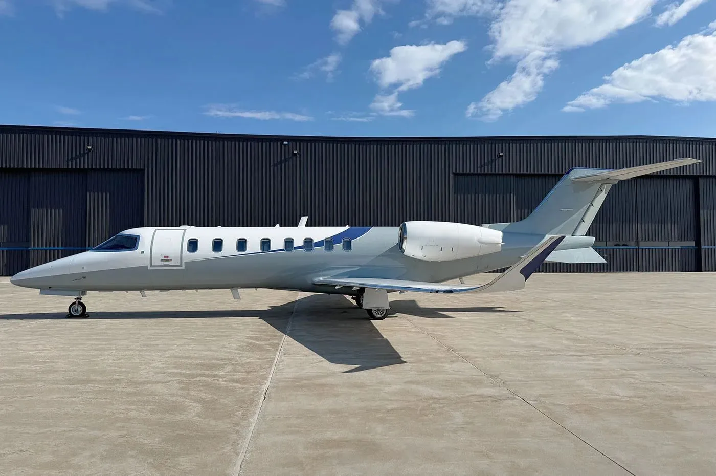 A Learjet 75 in a grey and white paint scheme with blue accents, parked on a tarmac in front of a dark hangar.