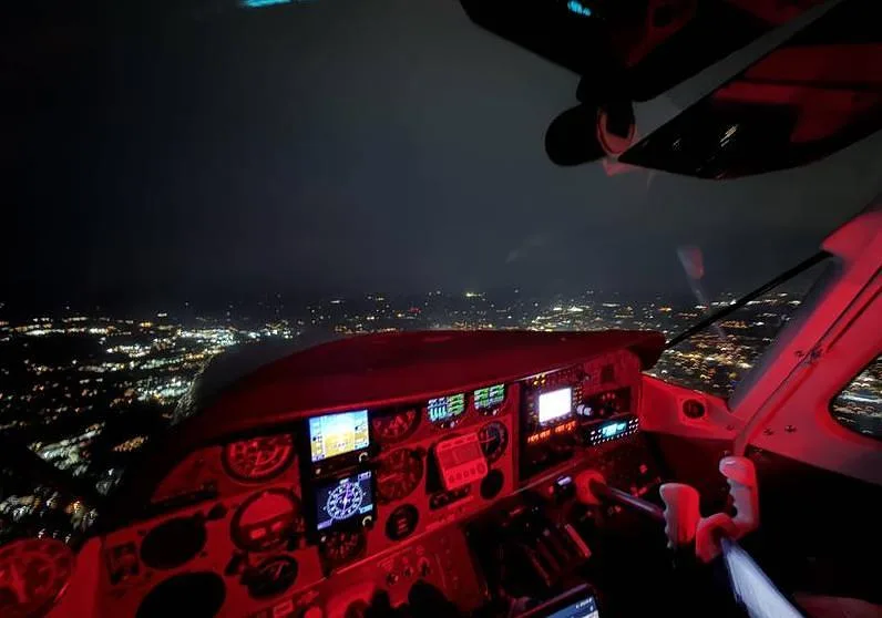 The aircraft's cockpit illuminated in red light during a night flight, with city lights visible below.