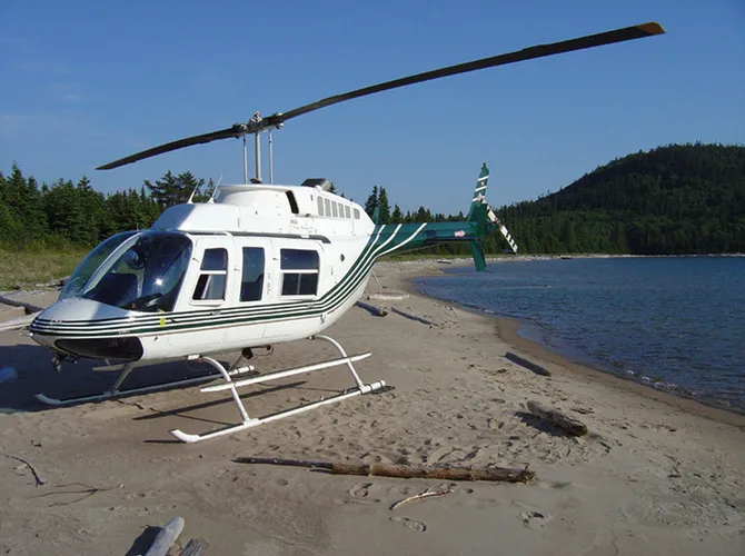 A white helicopter with green stripes is parked on a sandy beach next to a body of water with a forested hill in the background.