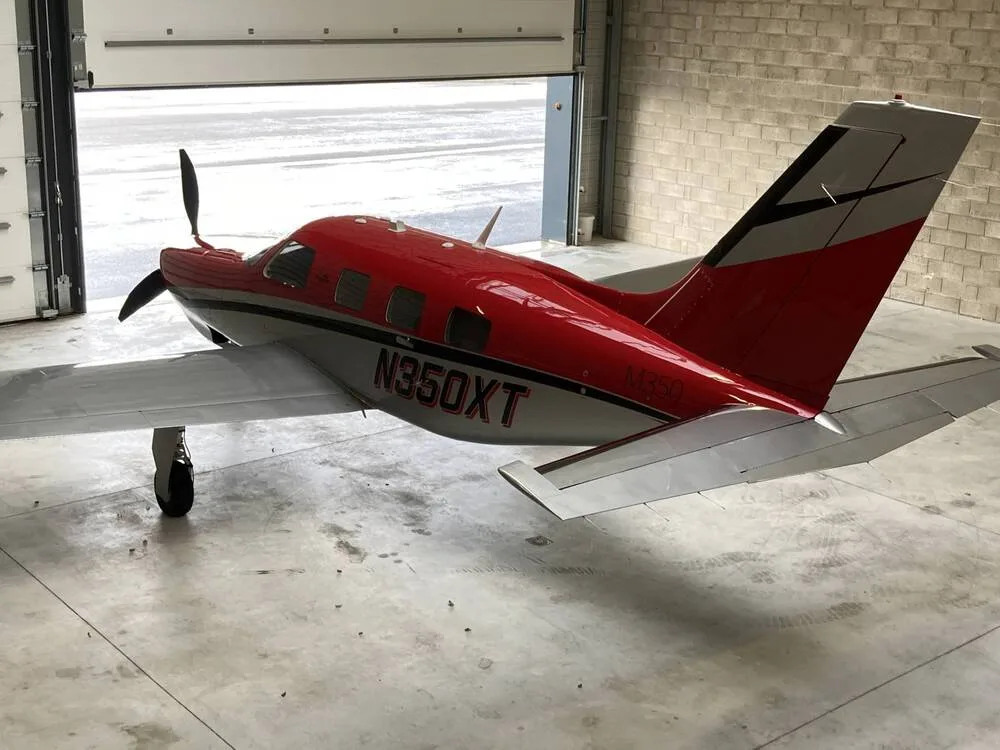 A rear three-quarter view of the red and silver Piper M350 aircraft, N350XT, partially in a hangar with the door open.