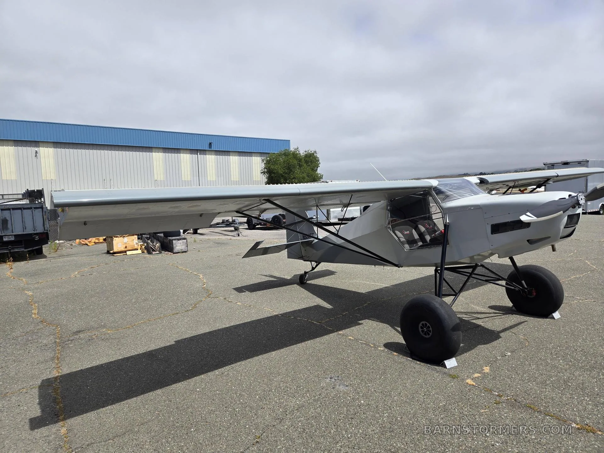 A light gray high-wing STOL aircraft with large tundra tires parked on an asphalt apron next to a hangar.