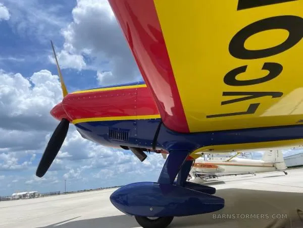 A close-up view of the red, blue, and yellow aircraft's propeller, engine cowling, and landing gear on the tarmac, with "LYCOMING" visible on the wing.
