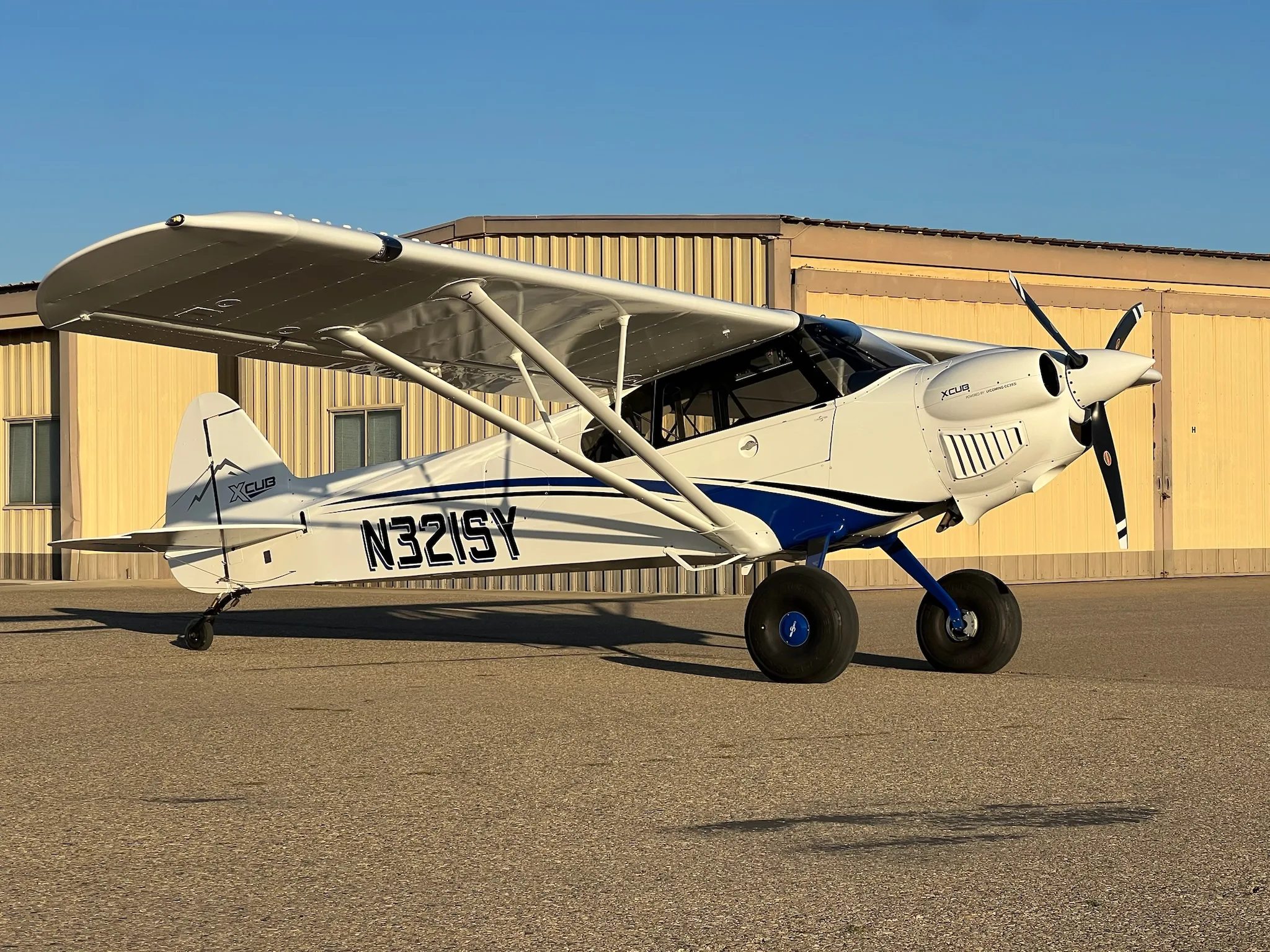 A white X-Cub aircraft with blue stripes and registration N321SY parked on an airfield with a building in the background.
