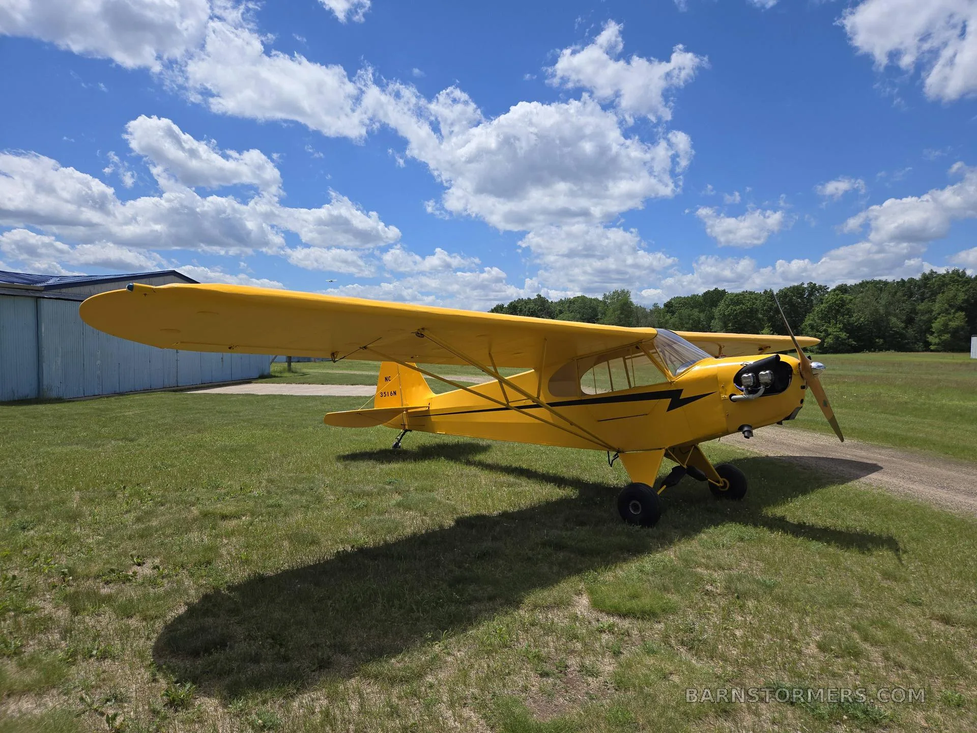 A yellow Piper Cub-style aircraft with black lightning bolt accents is parked on a grassy field under a blue sky.