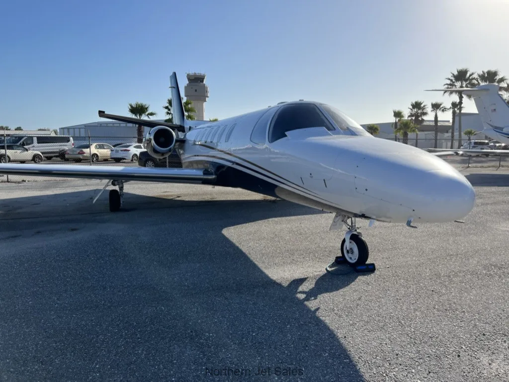Front side view of a white and black jet parked on the tarmac with palm trees and a control tower in the background.