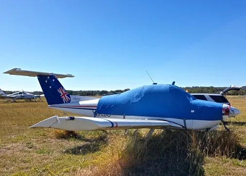 A white Beechcraft Skipper 77 with a blue cockpit cover and Australian flag tail, parked on a grassy field under a clear blue sky.