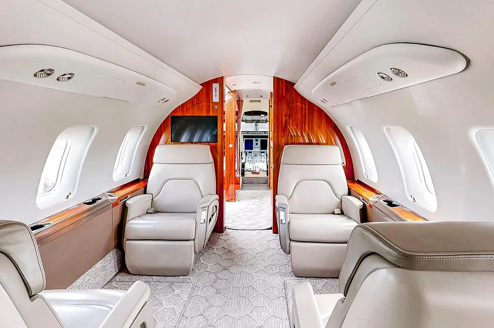 The luxurious cabin interior of a business jet features light-colored leather seats, wood paneling, and a patterned carpet.