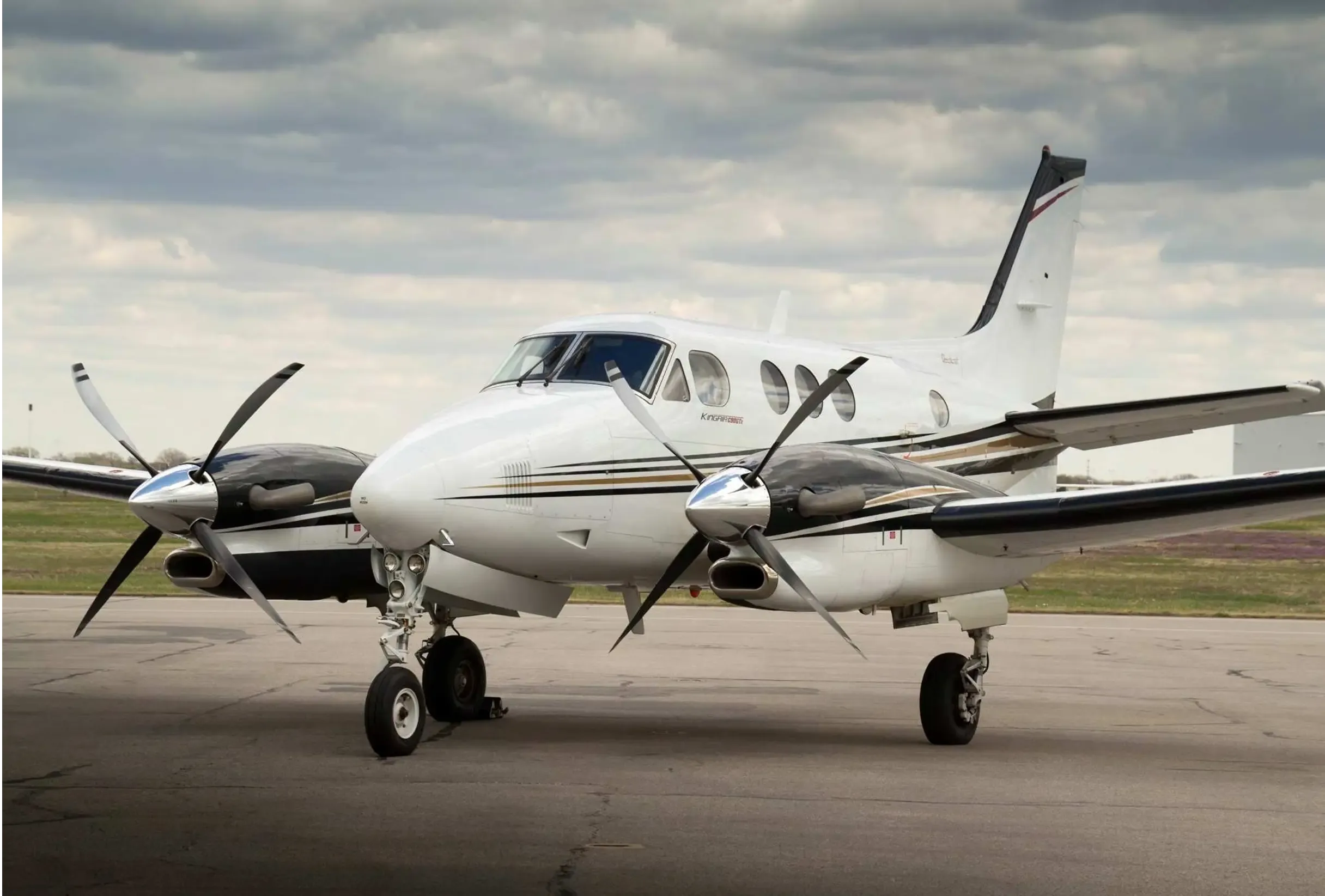 A white and black Beechcraft King Air 200 with gold stripes parked on a tarmac under a cloudy sky.