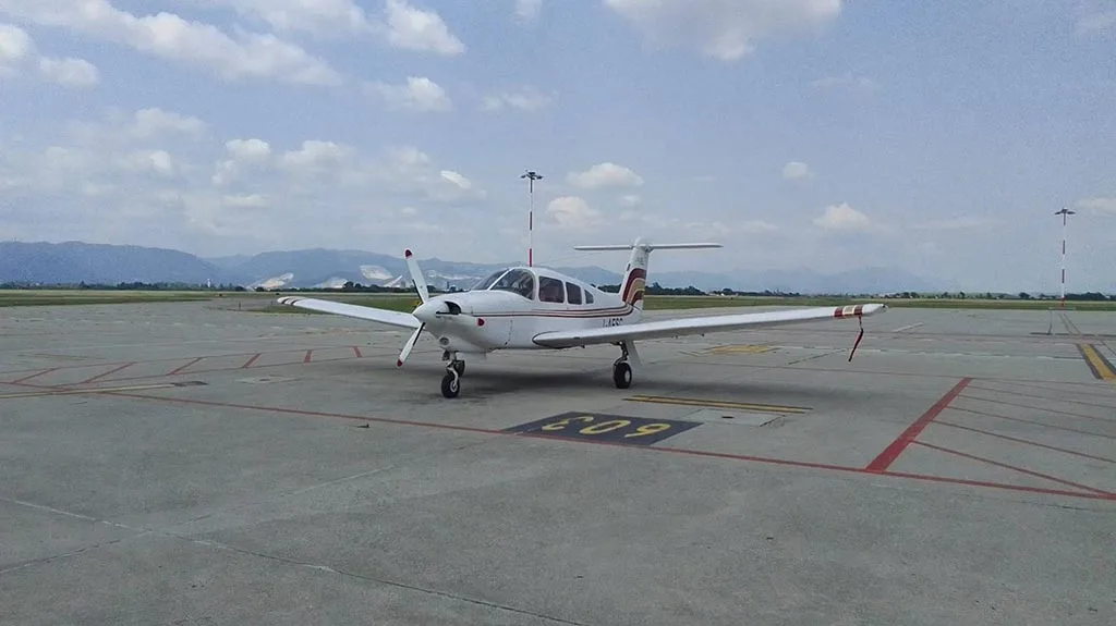 A white Piper PA-28 aircraft with red and gold stripes parked on an airfield with mountains in the background.