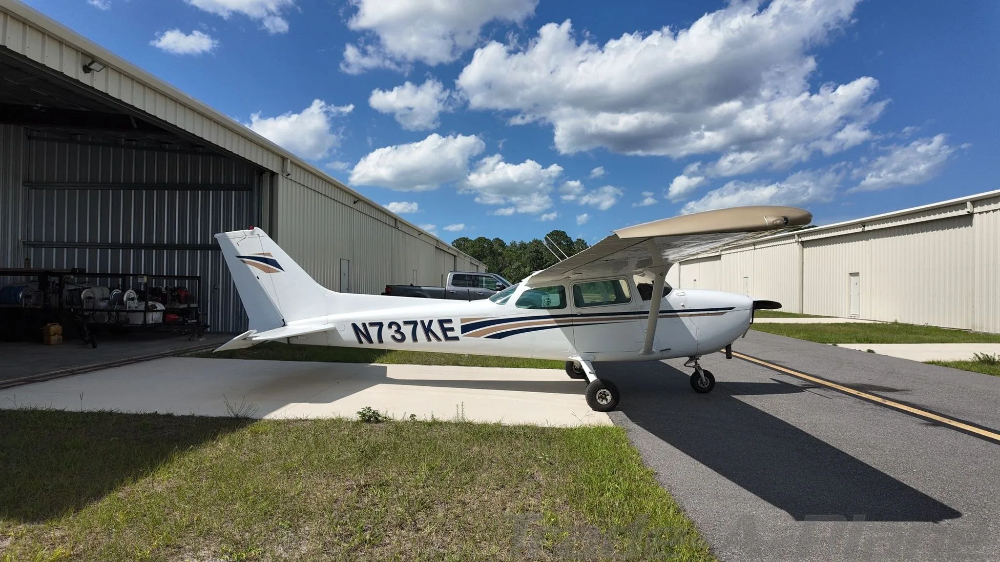 A white Cessna 172 Skyhawk with brown and blue stripes, tail number N737KE, parked outside a hangar on a sunny day.