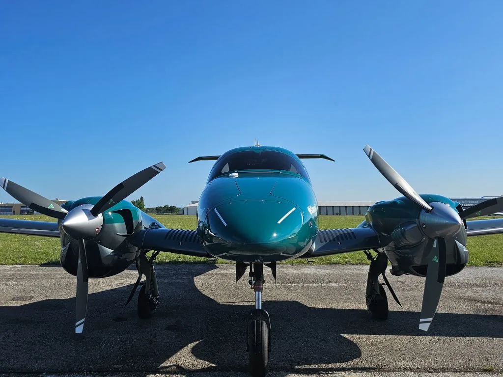 A dark green Diamond DA62 aircraft is parked on a tarmac, viewed from the front, under a clear blue sky.