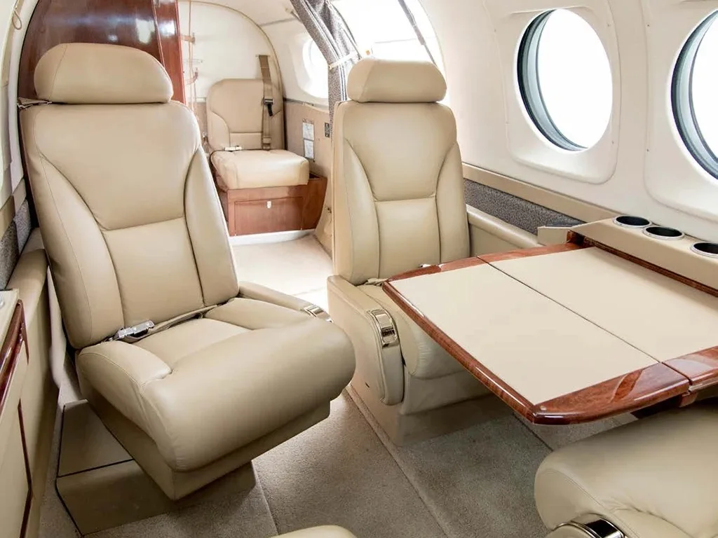 A section of an aircraft cabin interior featuring two beige leather seats and a fold-out wooden table with cup holders.