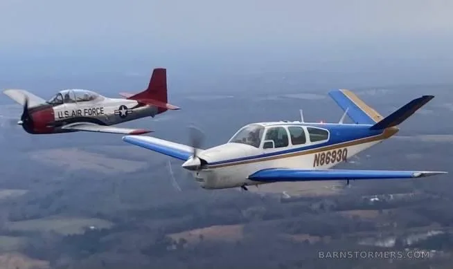 A white, blue, and gold Beechcraft Bonanza with tail number N8693Q flies alongside a red and silver U.S. Air Force T-28 Trojan against a cloudy sky.