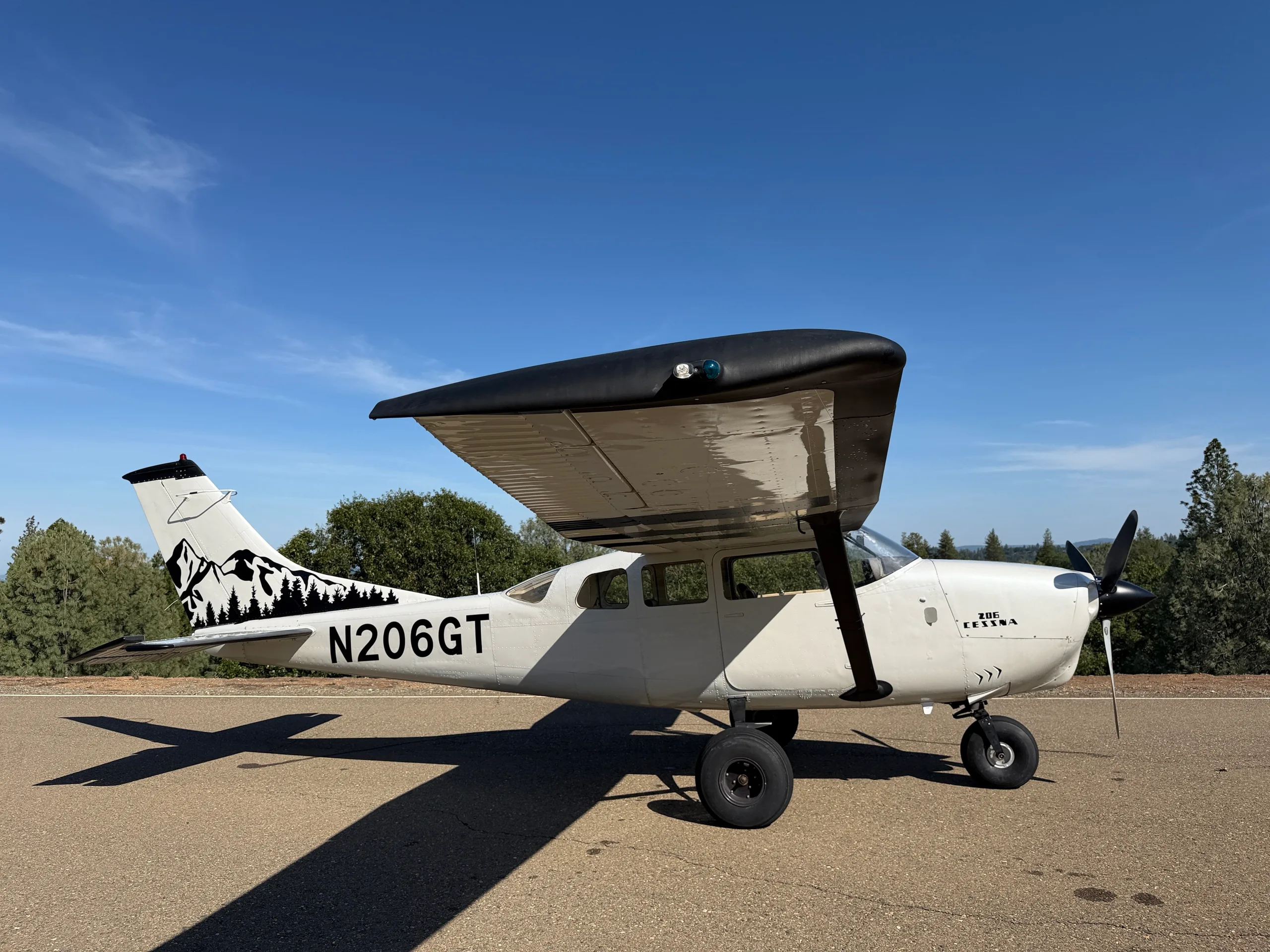 A white Cessna 206 with black wings and mountain tail art, parked on an asphalt strip with trees in the background.
