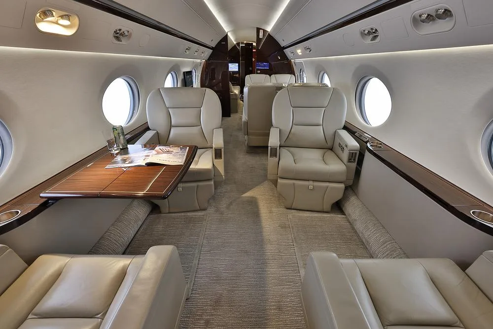 The luxurious cabin interior of a private jet, featuring beige leather seats, a polished wooden table, and large oval windows.