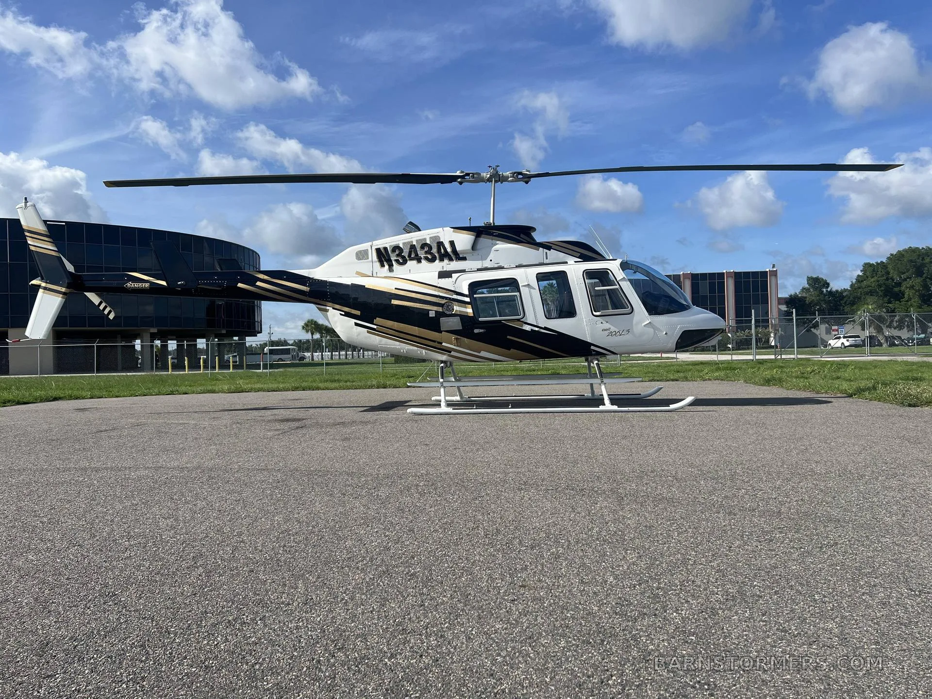 A white and gold Bell 206 helicopter (N343AL) is parked on an asphalt pad with buildings and a blue sky in the background.