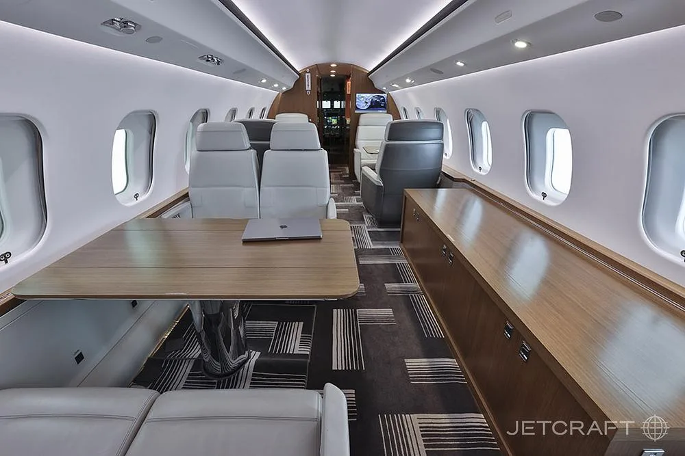 The aircraft cabin interior featuring a large wooden table, light grey seats, and a dark patterned carpet.
