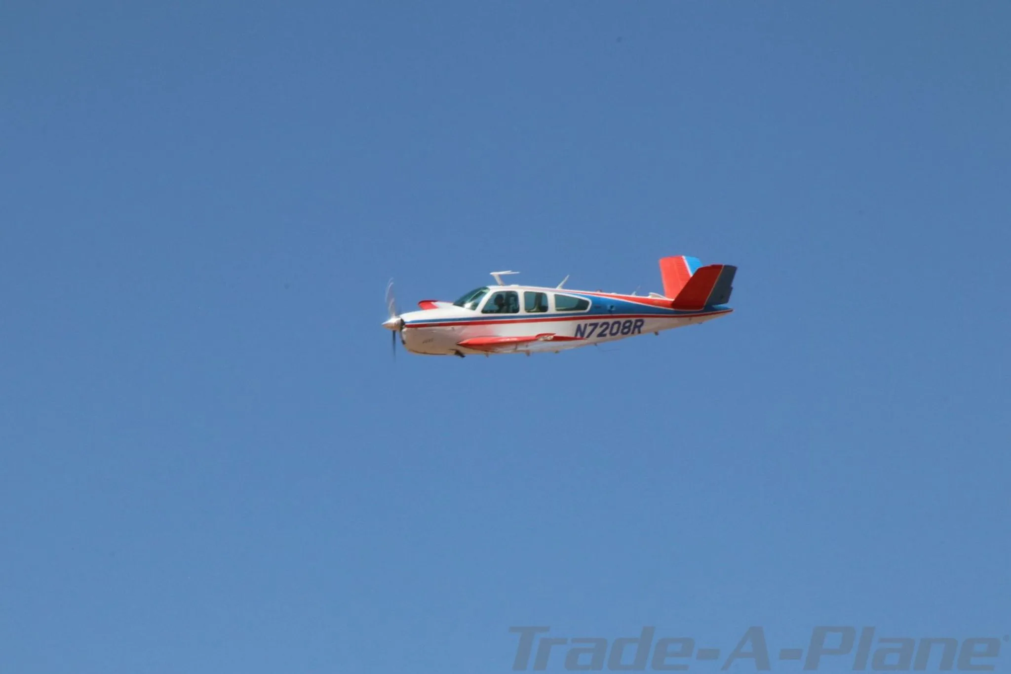 A white, red, and blue Beechcraft Bonanza N7208R is flying against a clear blue sky, viewed from the side.