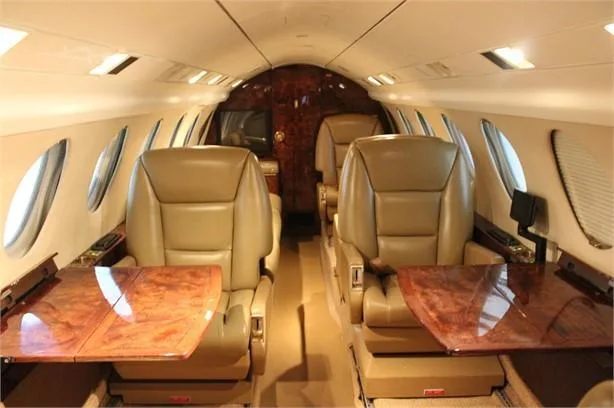 The luxurious aircraft cabin features tan leather seats, polished wood tables, and overhead lighting.