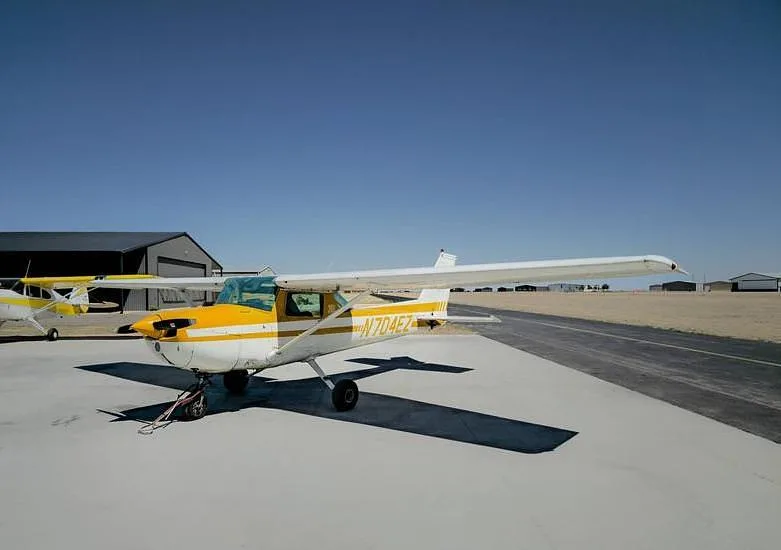 A white and yellow Cessna 150, registration N704EZ, parked on a tarmac with hangars in the background.