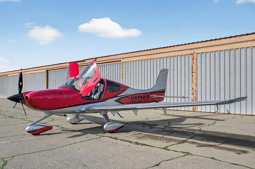 A red and silver single-engine aircraft, N512KP, with its gull-wing doors open, parked on an airfield.