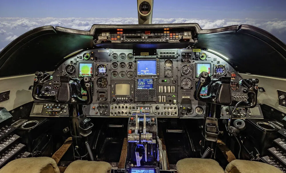 A detailed view of the Learjet 45 cockpit with multiple screens, gauges, and control yokes, showing a sky view through the windshield.