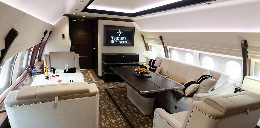 A luxurious aircraft lounge area with a large sofa, dining table, and a television displaying 'The Jet Business' logo.