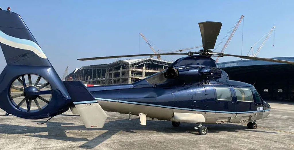 A rear-side view of a dark blue and silver Airbus H155 helicopter parked on an airfield with construction cranes in the background.