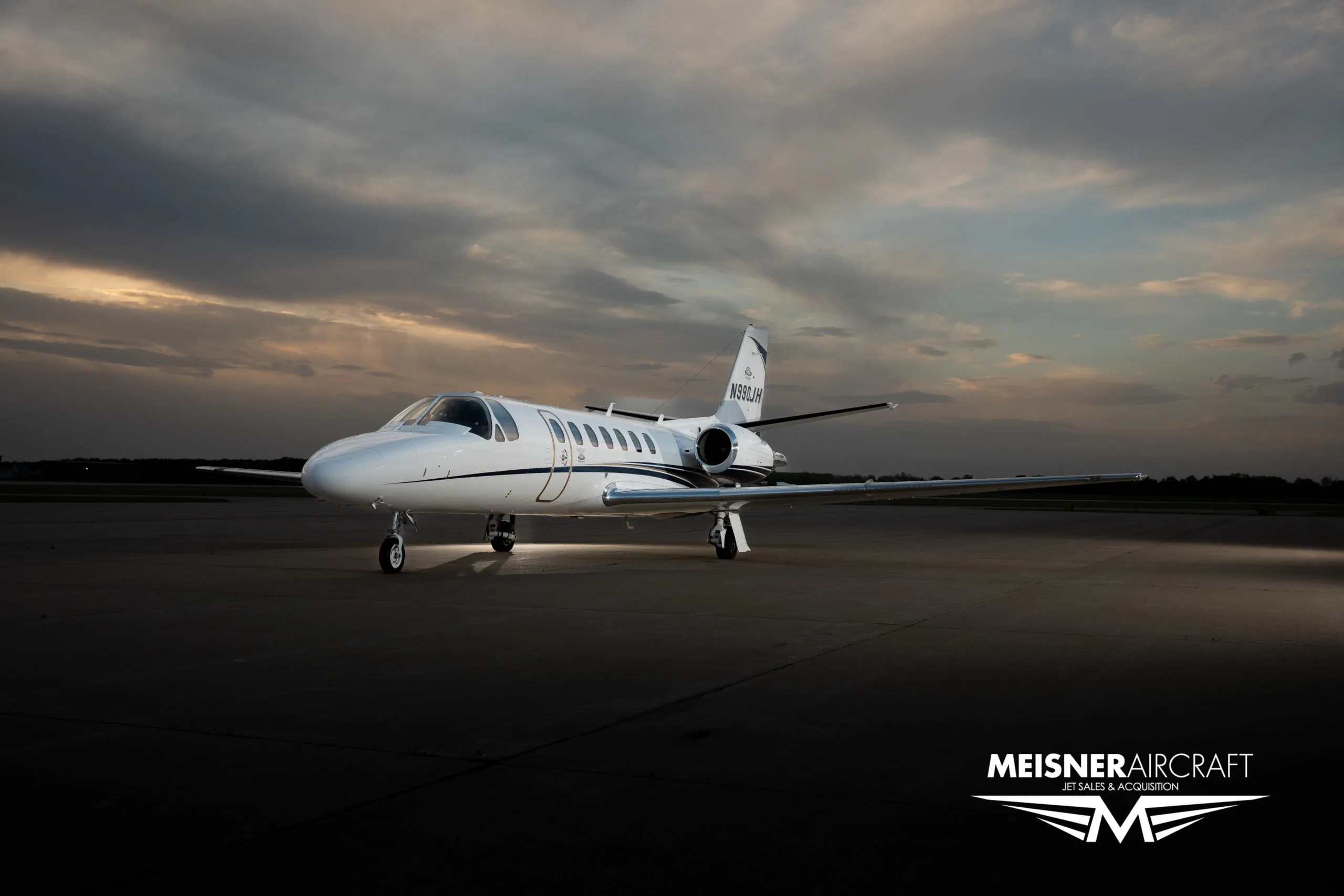 A white Cessna Citation jet with blue and black stripes, registration N990JH, parked on an airfield at sunset, with a 'Meisner Aircraft' logo.