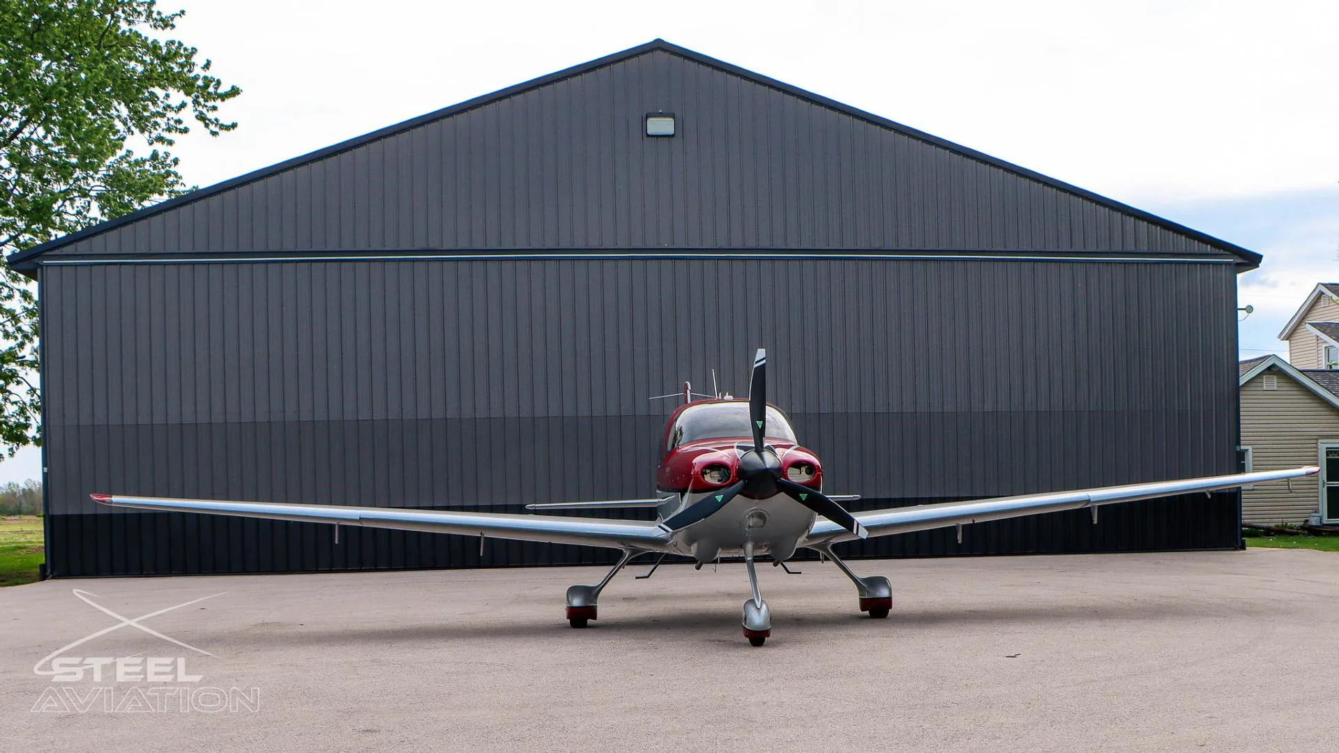 A red and white Cirrus SR22 aircraft is parked on an asphalt apron in front of a dark gray hangar.