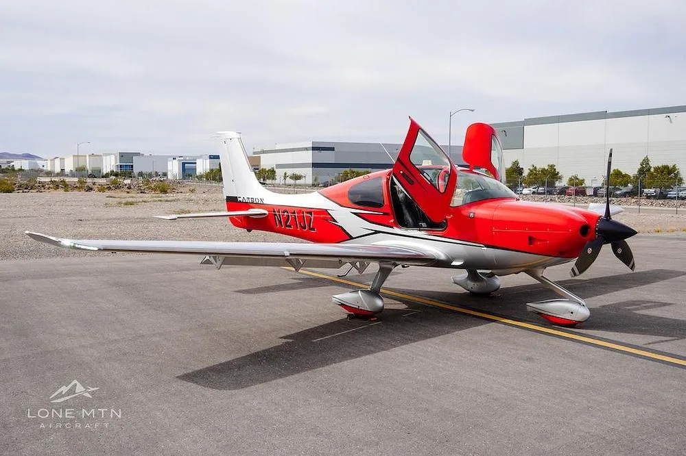 A red and silver Cirrus SR22 Carbon aircraft with its doors open, parked on an asphalt tarmac with buildings in the background.
