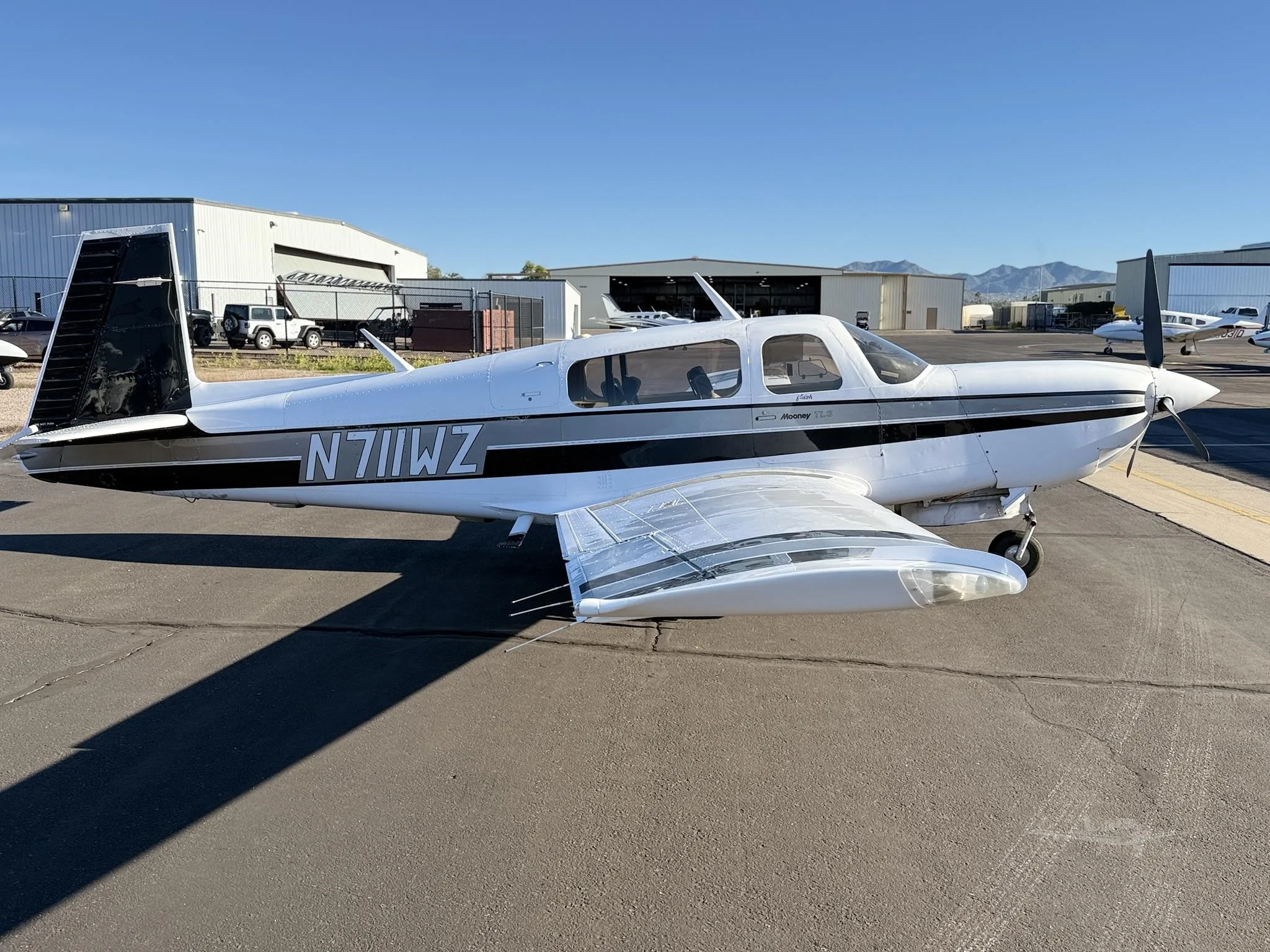 A white Mooney TLS aircraft with black and grey stripes is parked on an asphalt tarmac, with hangars and mountains in the background under a clear blue sky.