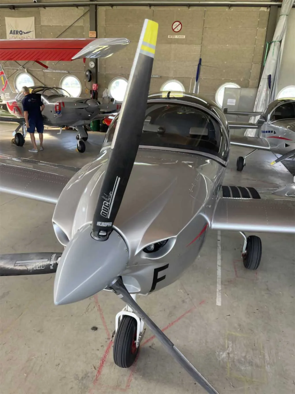 Front view of a silver Sonaca aircraft with a black propeller, parked in a hangar.
