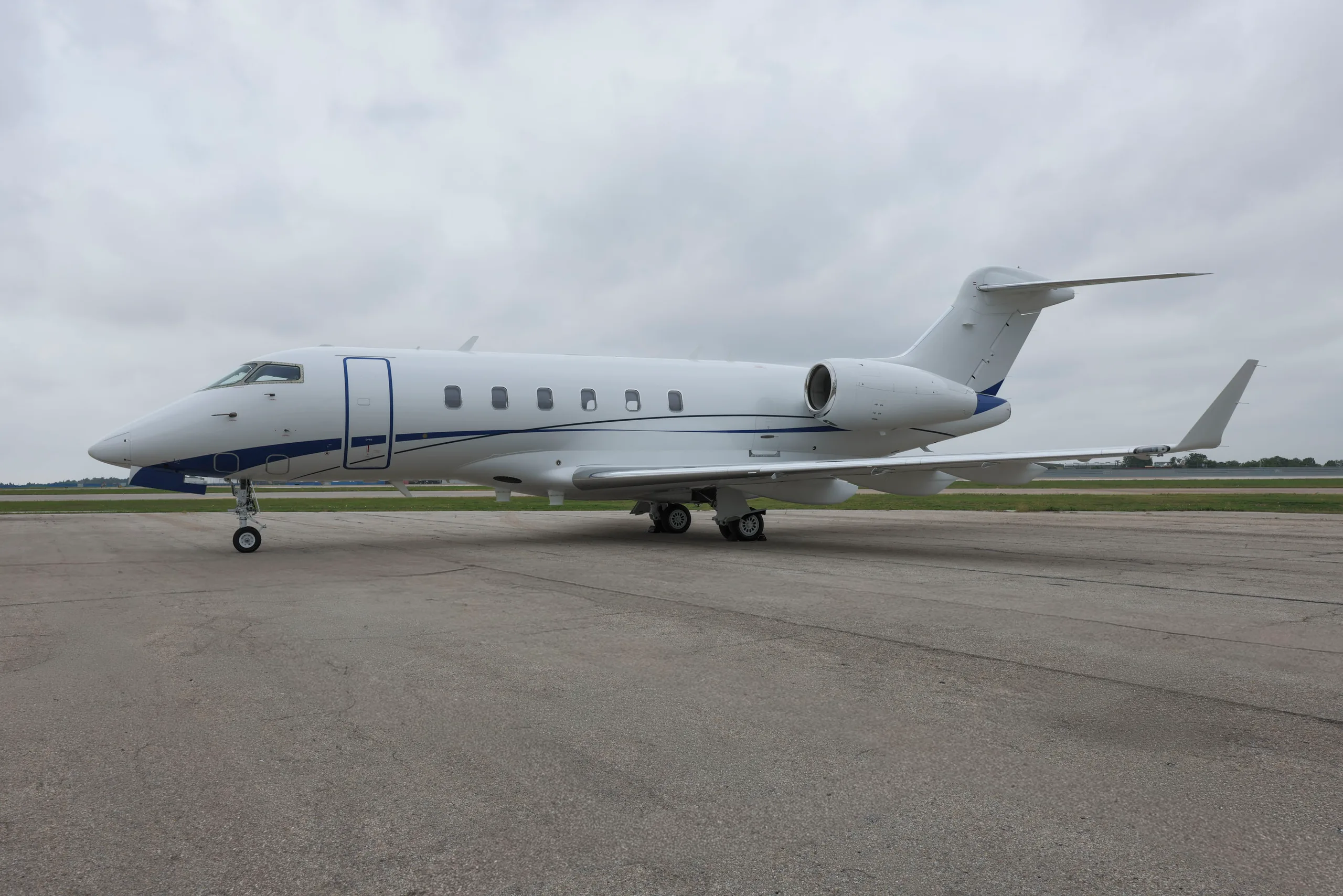 A white business jet with blue accent stripes is parked on an asphalt tarmac under a cloudy sky.