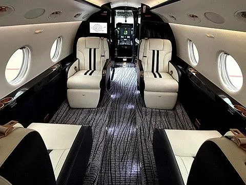 The luxurious interior cabin of a private jet, featuring cream and black seats, patterned carpet, and a view into the cockpit.