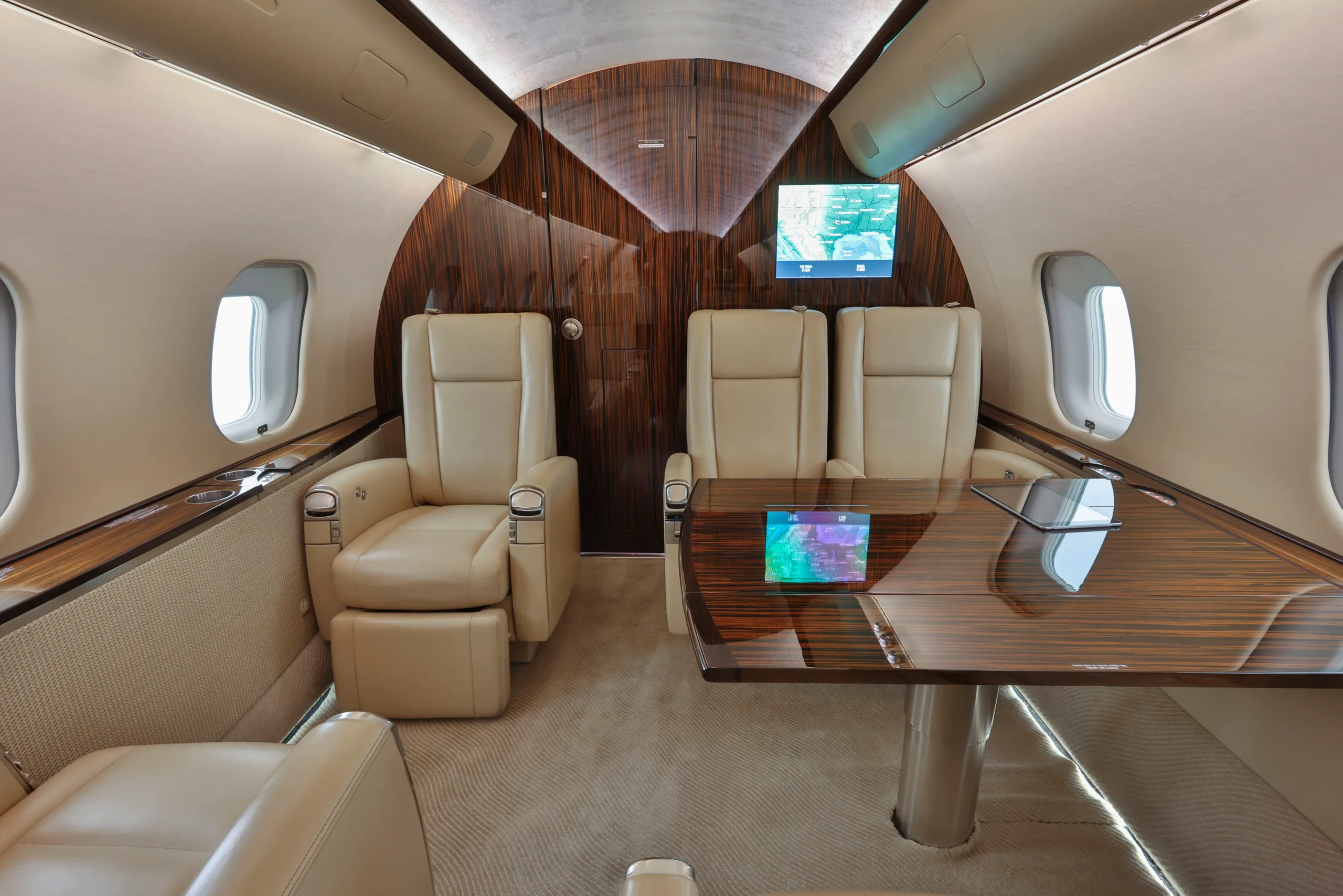 A section of the business jet's cabin with two beige leather seats, a wooden table, and two wall-mounted screens.