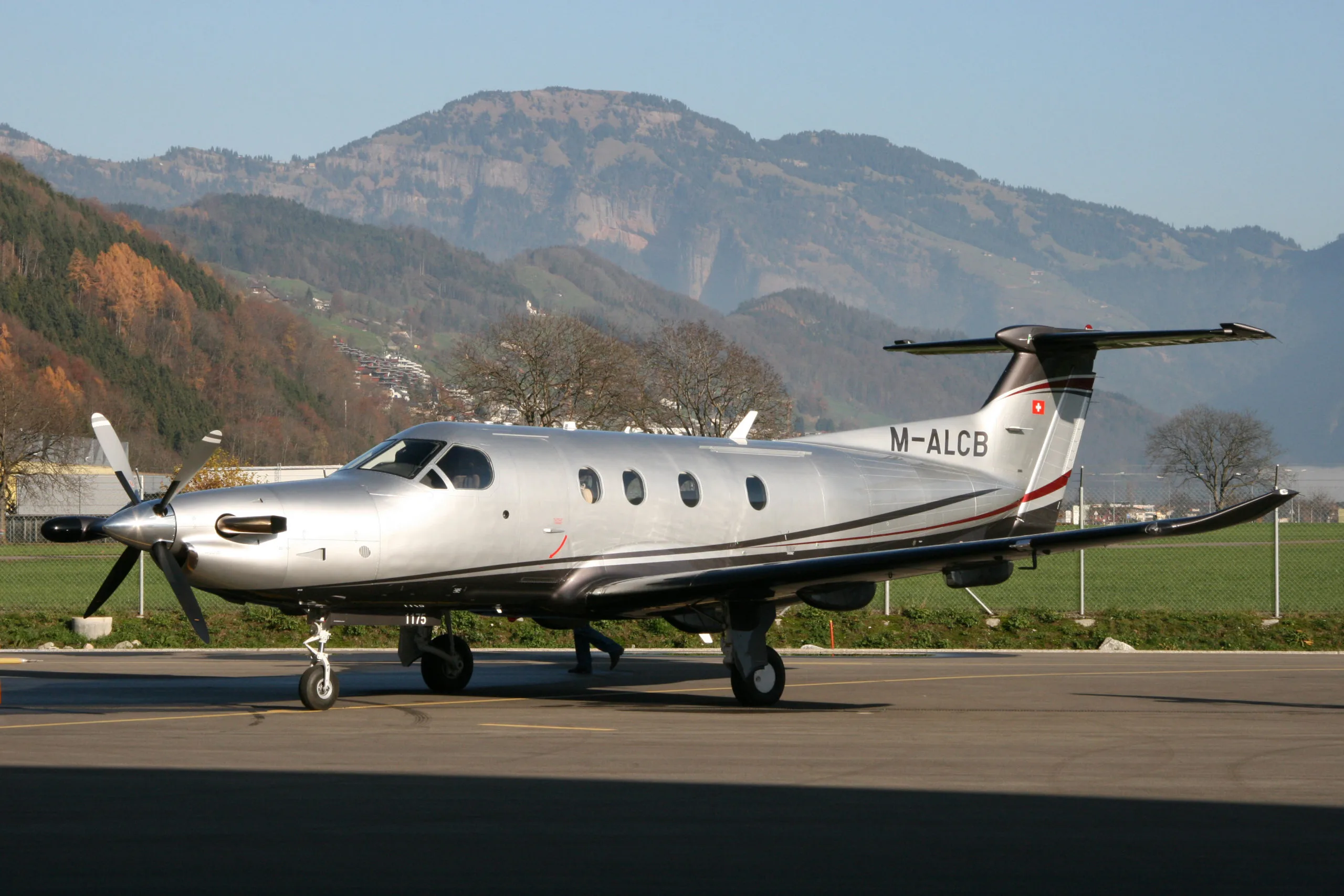 A silver Pilatus PC-12 aircraft with dark grey and red stripes is parked on an airfield with mountains in the background.