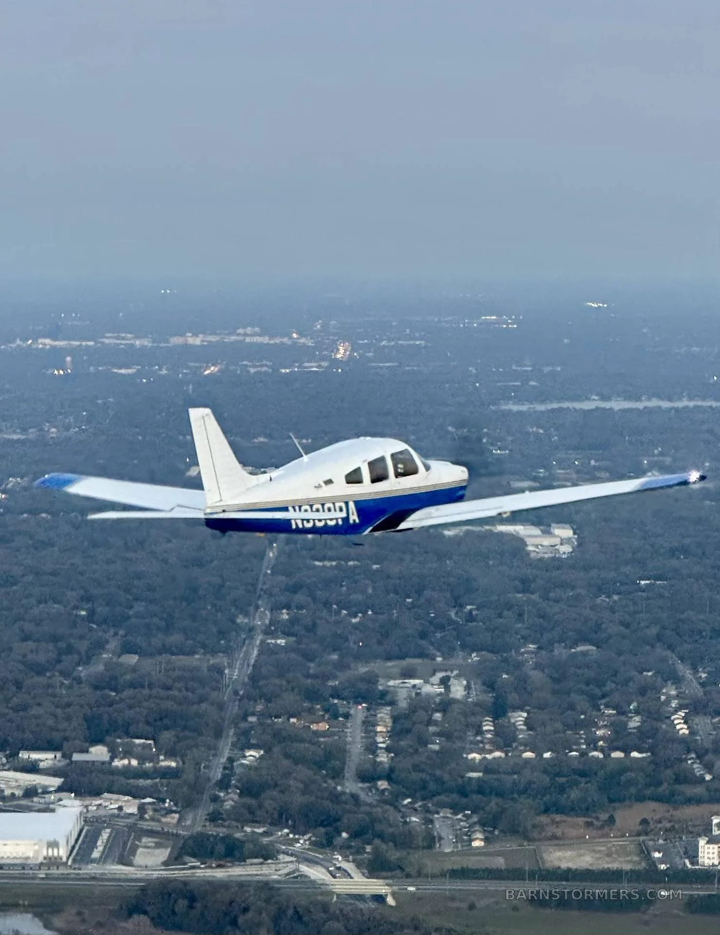 A white and blue Piper Cherokee aircraft (N338PA) is flying over a tree-covered suburban landscape at dusk.