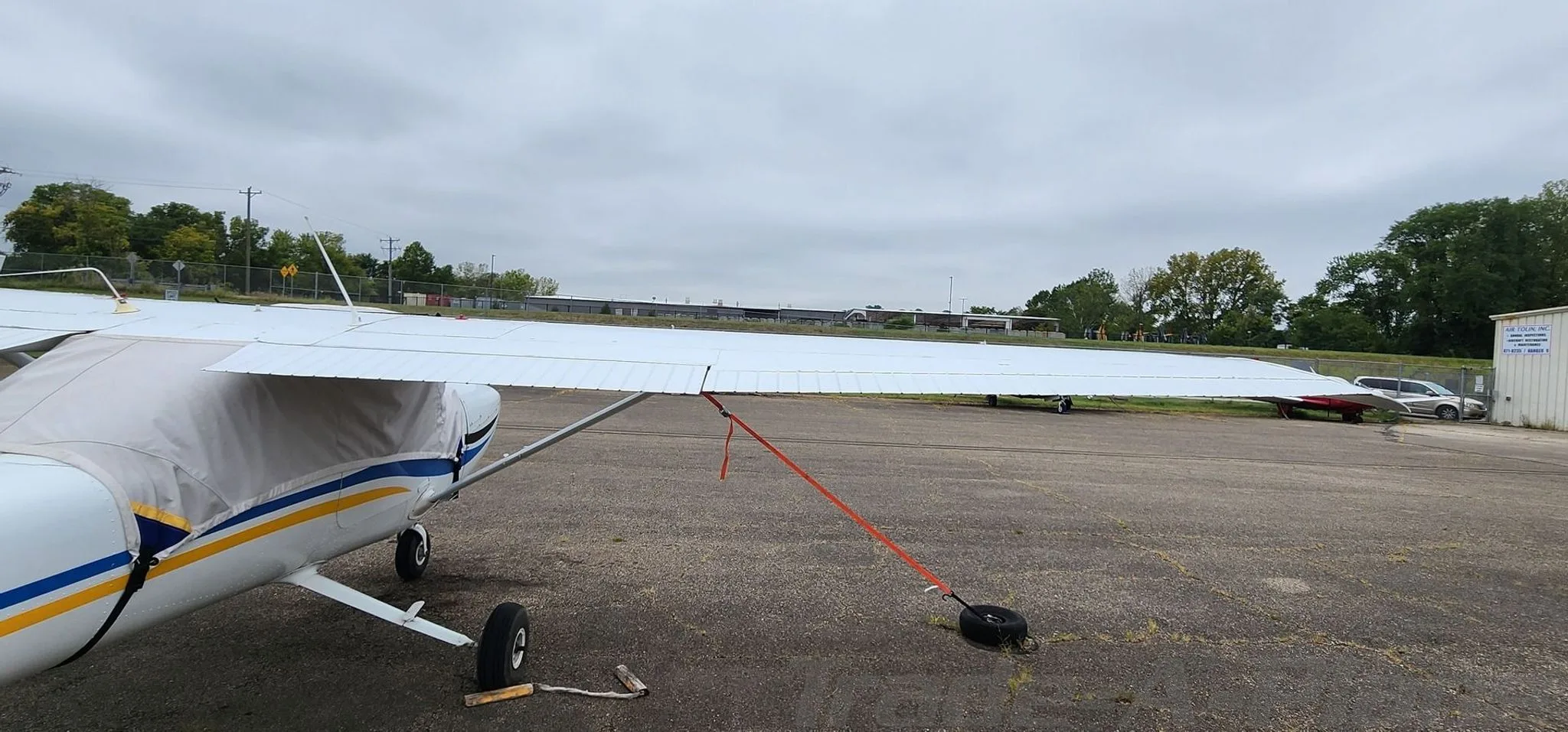 A side view of the white aircraft's wing and fuselage, showing it tied down on the tarmac next to a hangar.