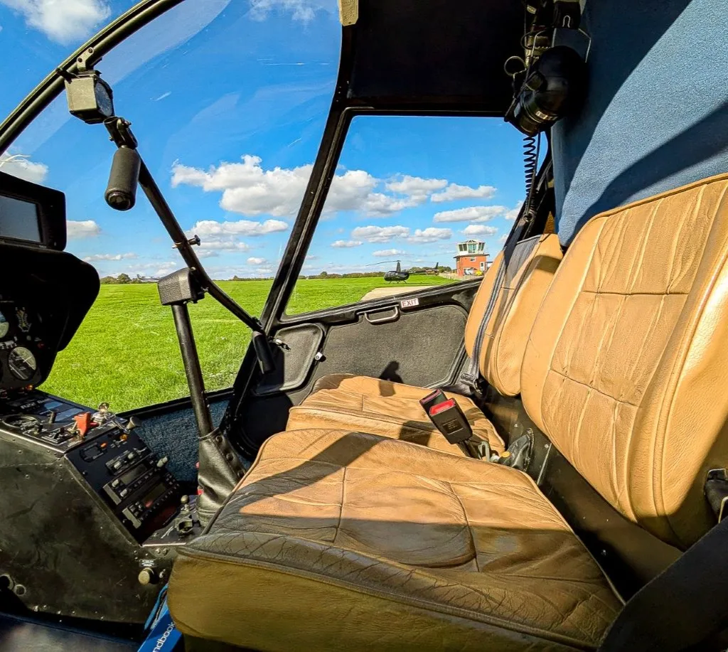 The tan leather interior seats of a helicopter with a view of a grassy airfield and a control tower through the windshield.