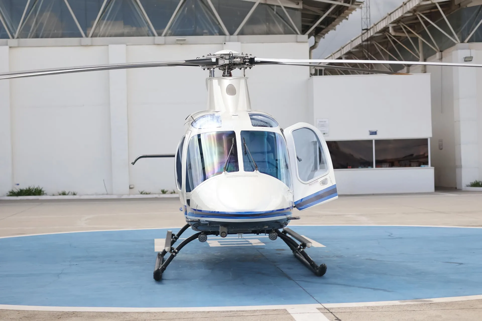 A front view of a white and blue helicopter with its doors open, parked on a blue helipad.