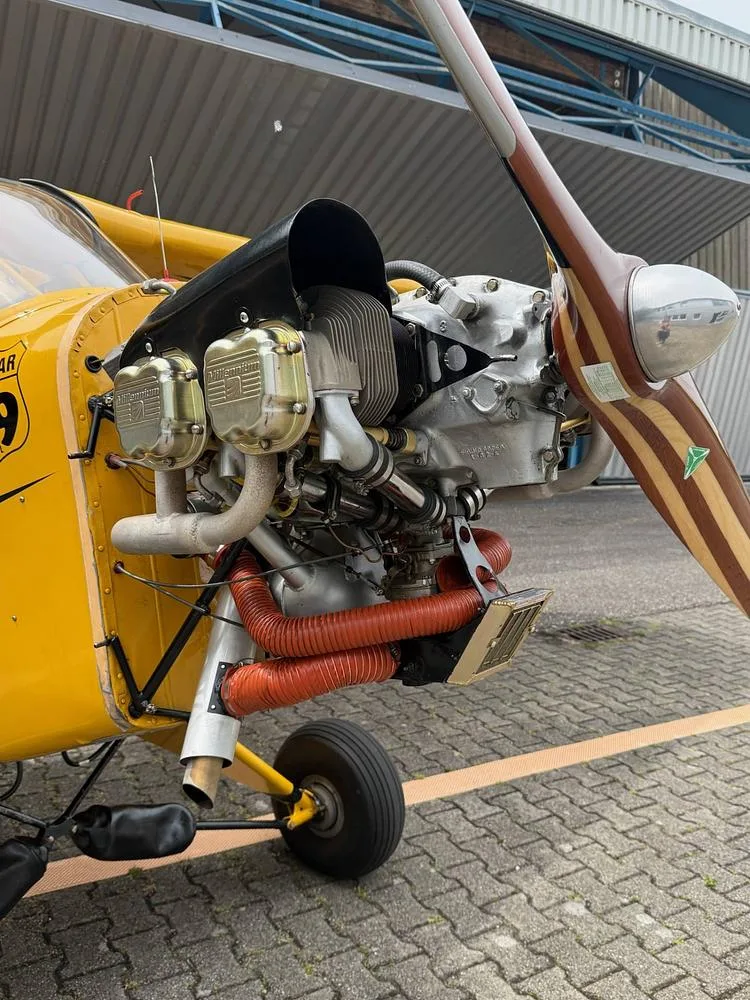 Close-up view of the exposed engine and wooden propeller of a yellow aircraft parked on an airfield.