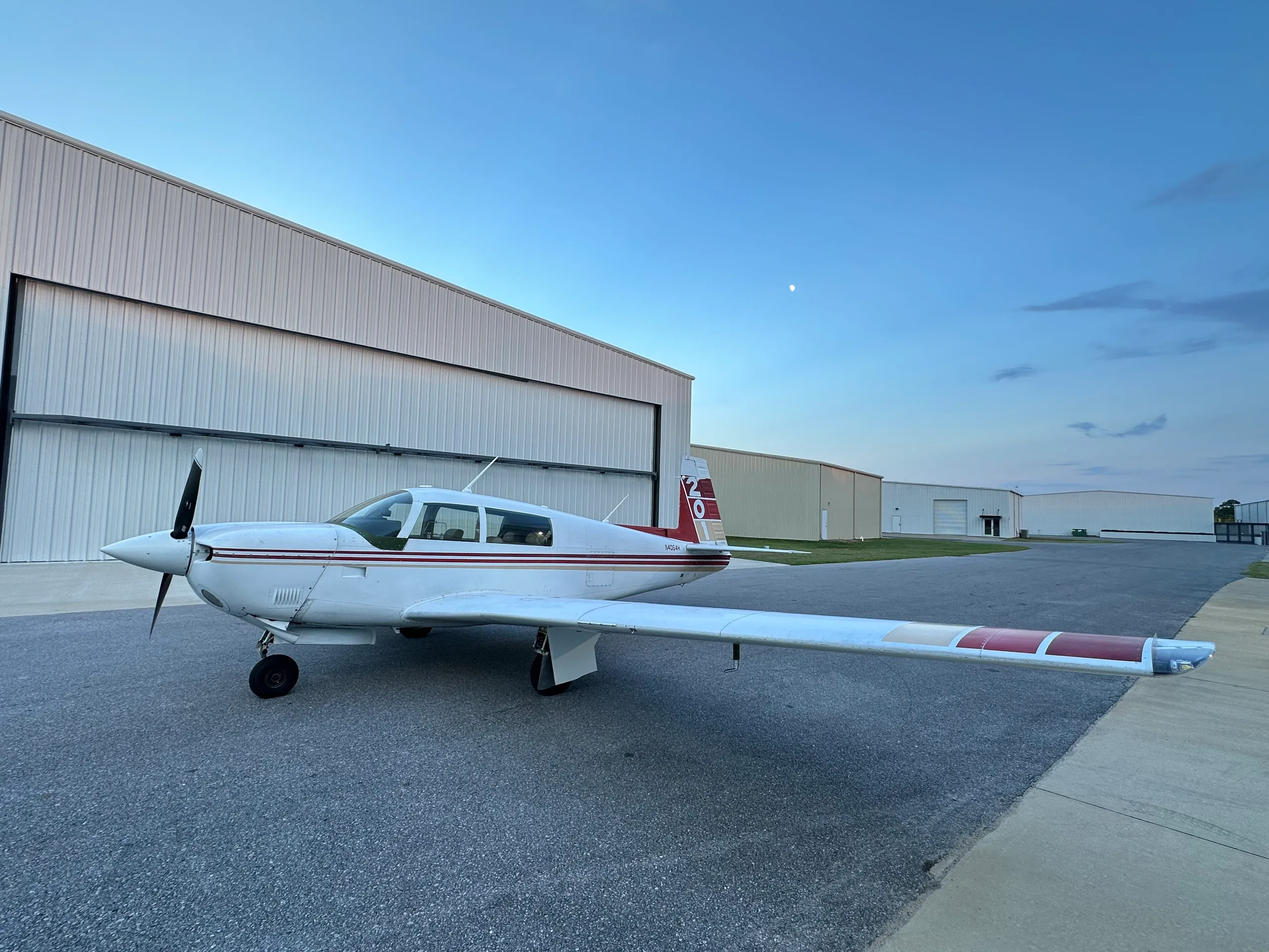 A white single-engine aircraft with red and gold stripes on the tail and wings, parked on an asphalt apron outside hangars with a moon in the sky.