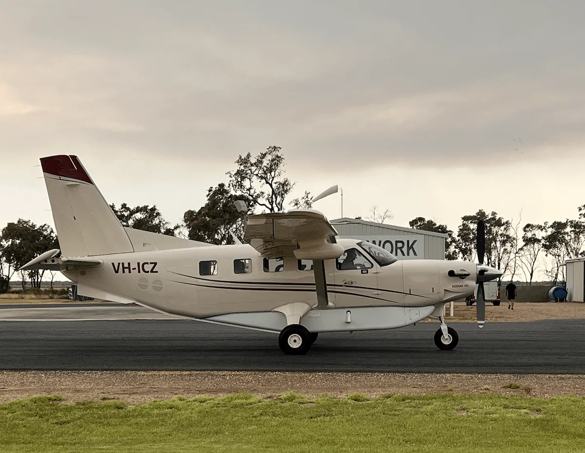 A beige Quest Kodiak 100 aircraft with red stripes, registration VH-ICZ, parked on an airfield under a cloudy sky.