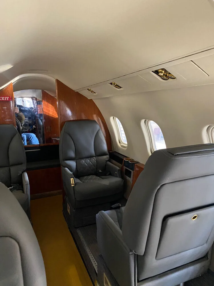 A view inside the business jet's cabin, showing dark grey leather seats and wood accents along the walls.