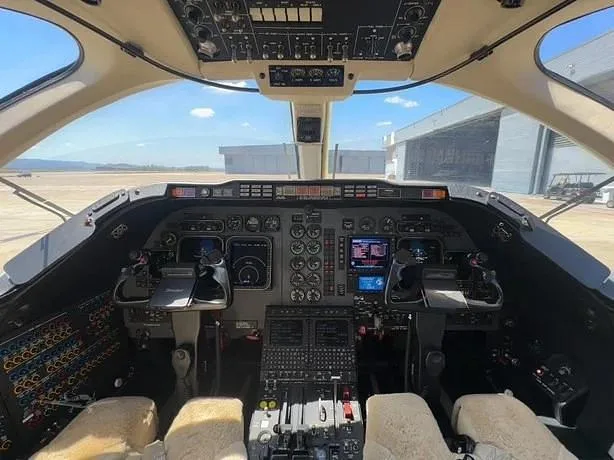 The cockpit of a private jet features a complex instrument panel with multiple screens, controls, and a view of the tarmac outside.