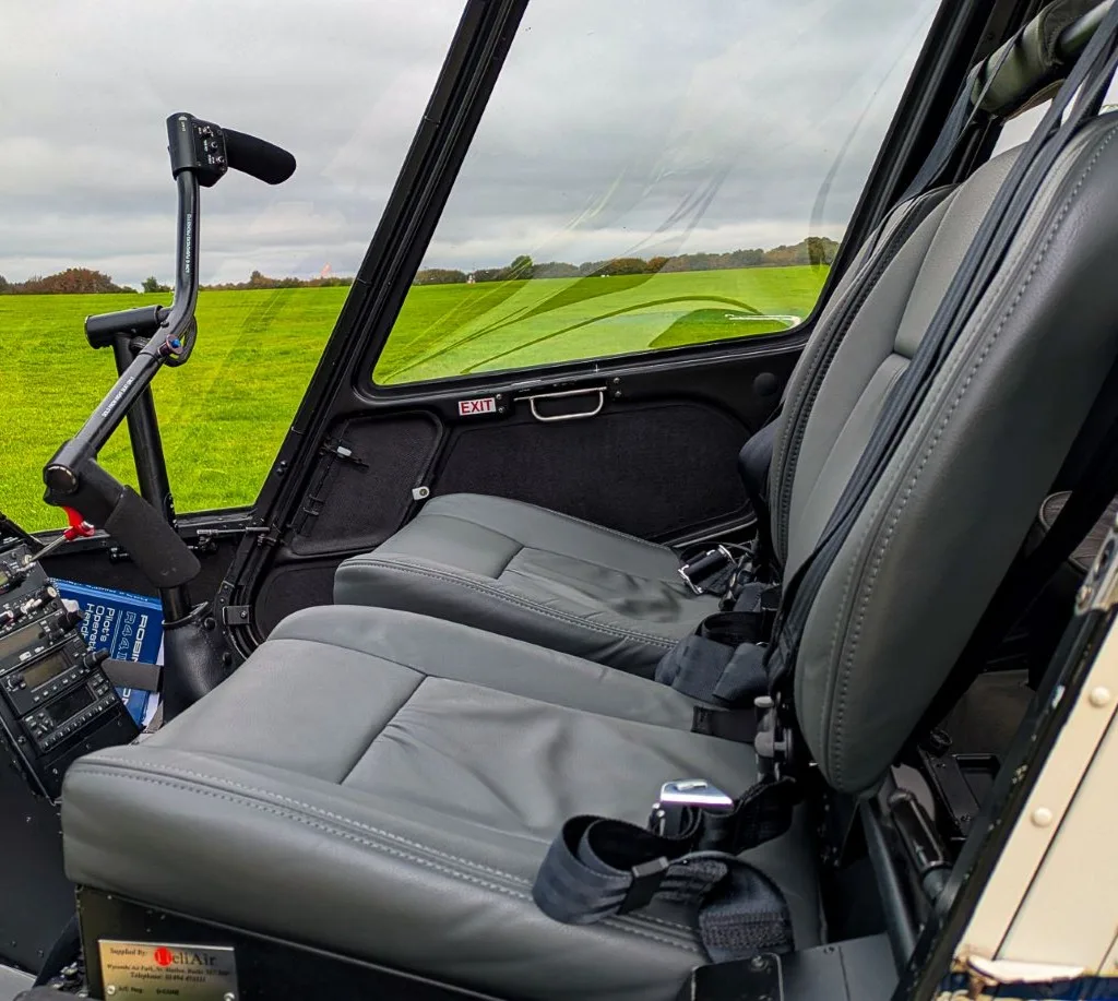 The grey leather interior seats and controls of a helicopter, with a view of a green field through the windows.
