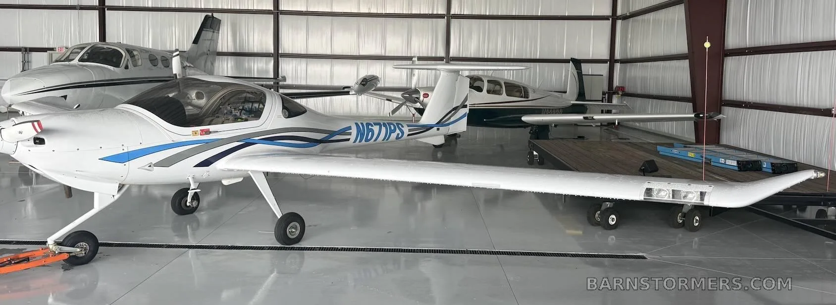 A white Diamond DA20-C1 Eclipse aircraft with blue and grey stripes, registration N671PS, parked inside a hangar with other planes.