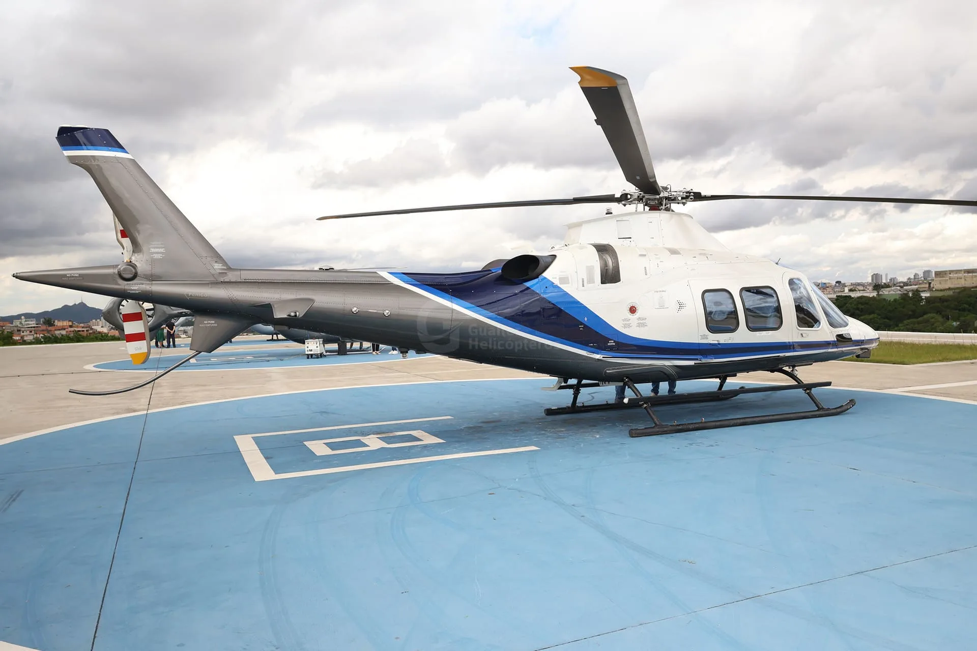 A white and blue helicopter with a grey tail is parked on a blue helipad under a cloudy sky.
