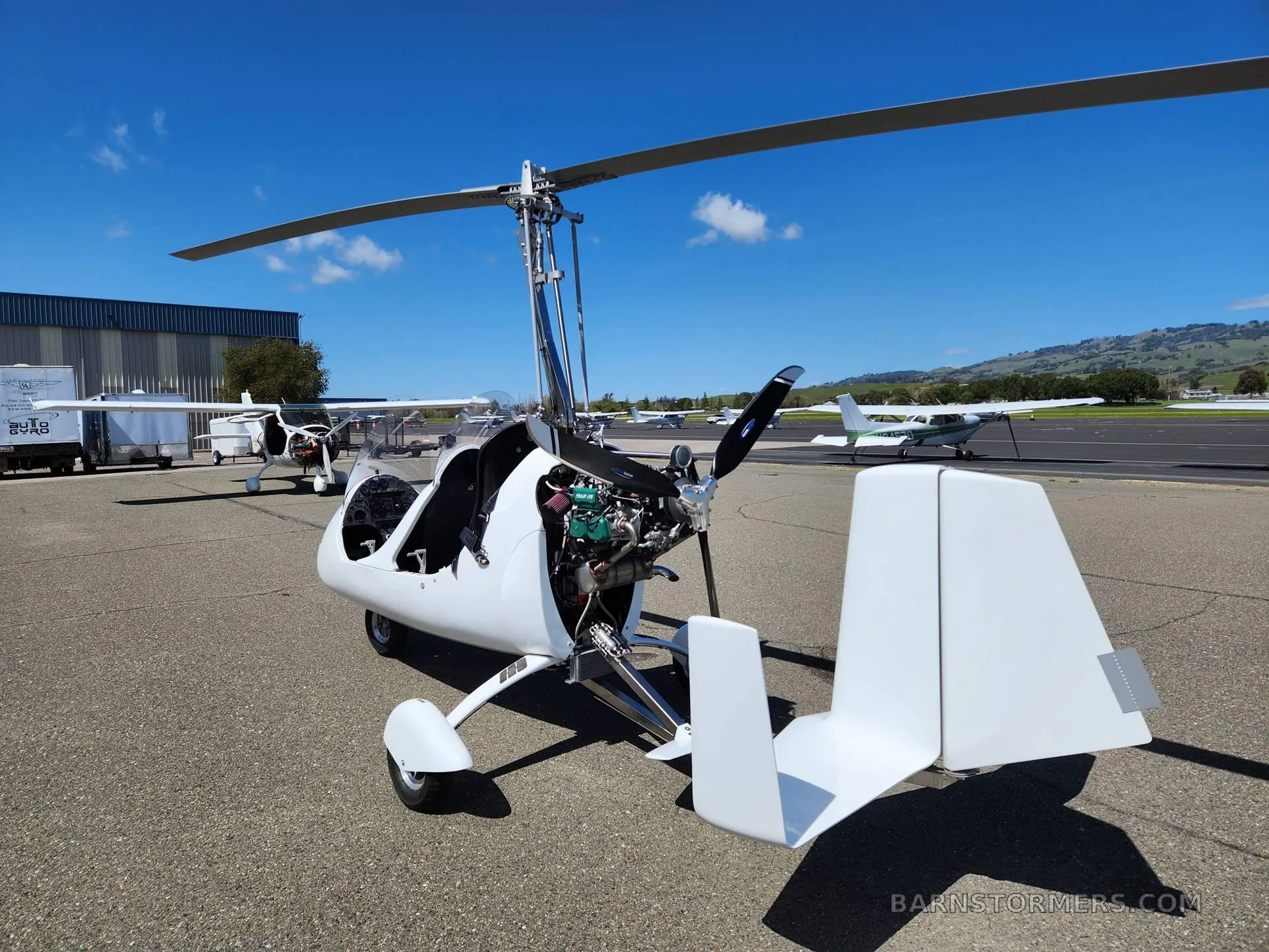 A white gyrocopter with its engine exposed, parked on an airfield tarmac under a clear blue sky.